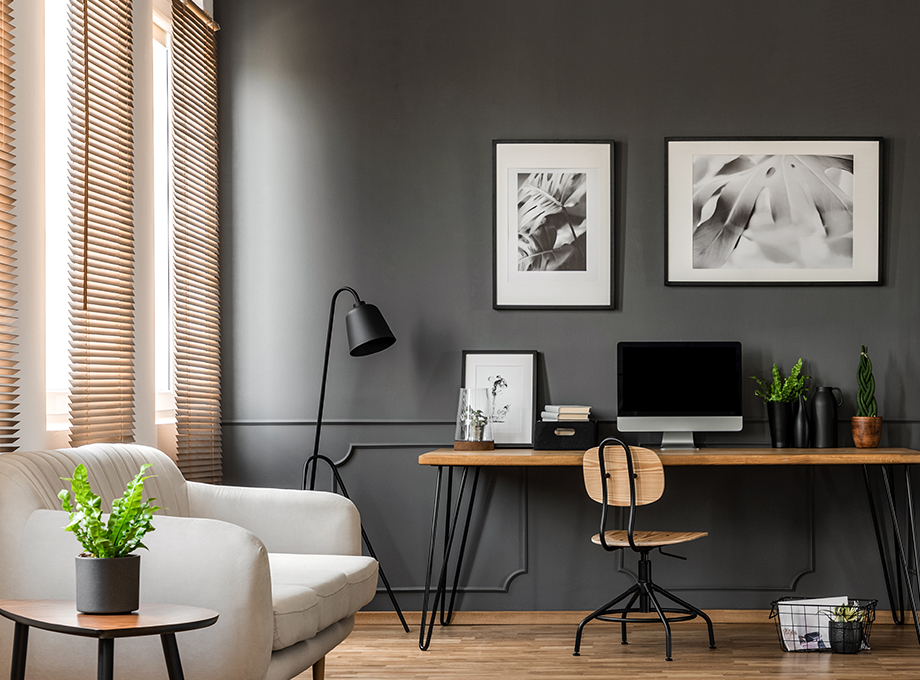 Modern home office with gray walls, wood blinds, a sleek desk, green plants, and minimalist black-and-white framed artwork.
