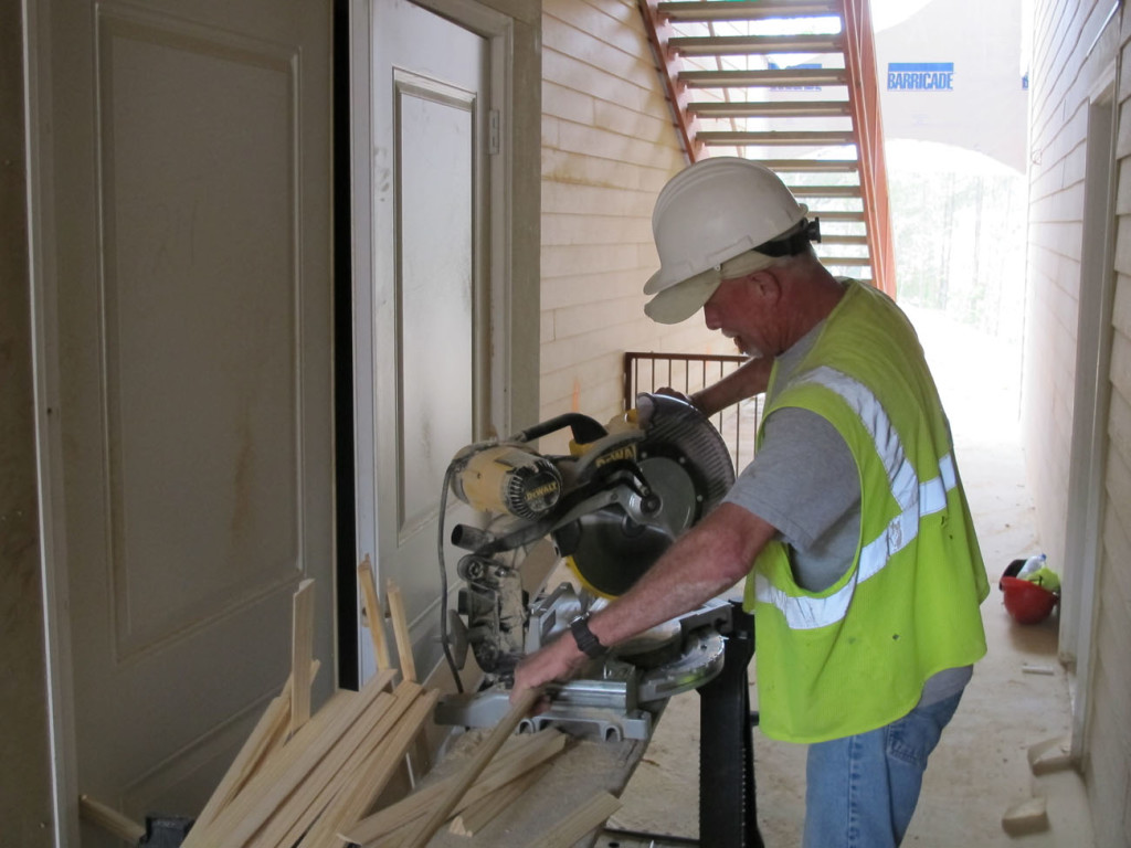 A construction worker wearing a hard hat and safety vest cuts wooden trim pieces using a miter saw at a multifamily building site.